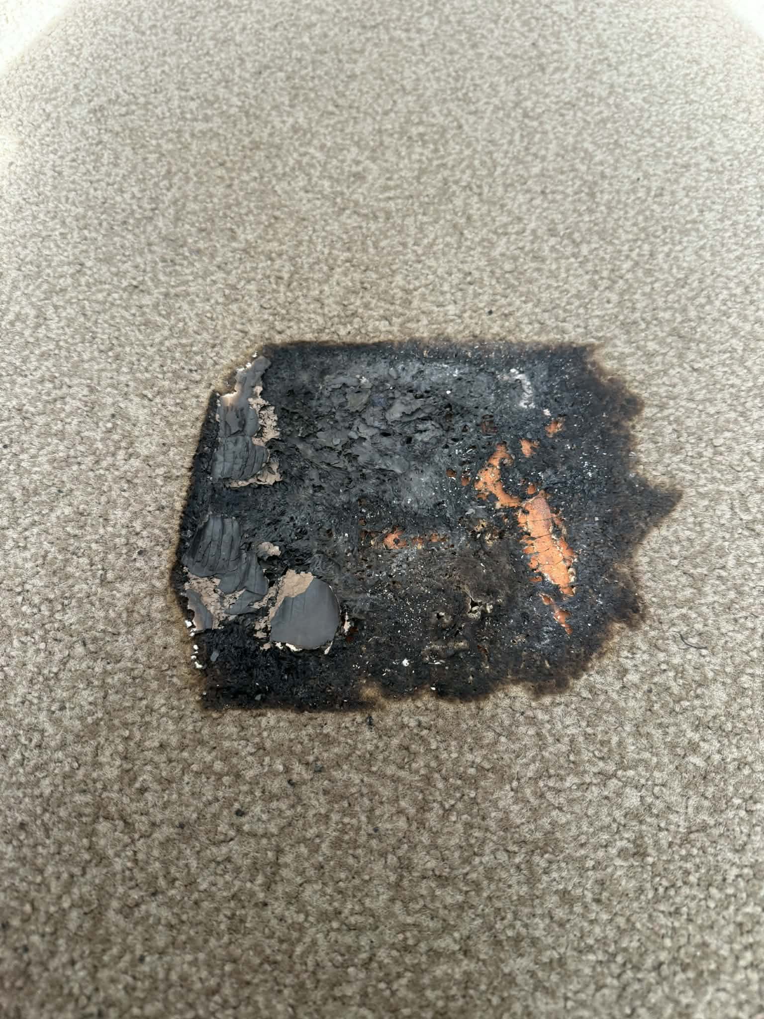 Beige carpet with a large charred and melted burn hole exposing the backing